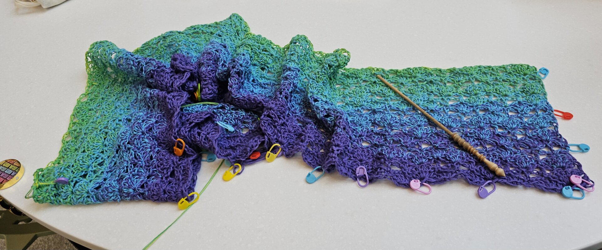 A long crocheted rectangle bunched up on a white table with many multicolored stitch markers along the edge, and a birch crochet hook along the top. The piece of fabric is lacy, starting with dark purple on the bottom edge, slowly changing to blue and then green with the tiniest hint of yellow on the top row.