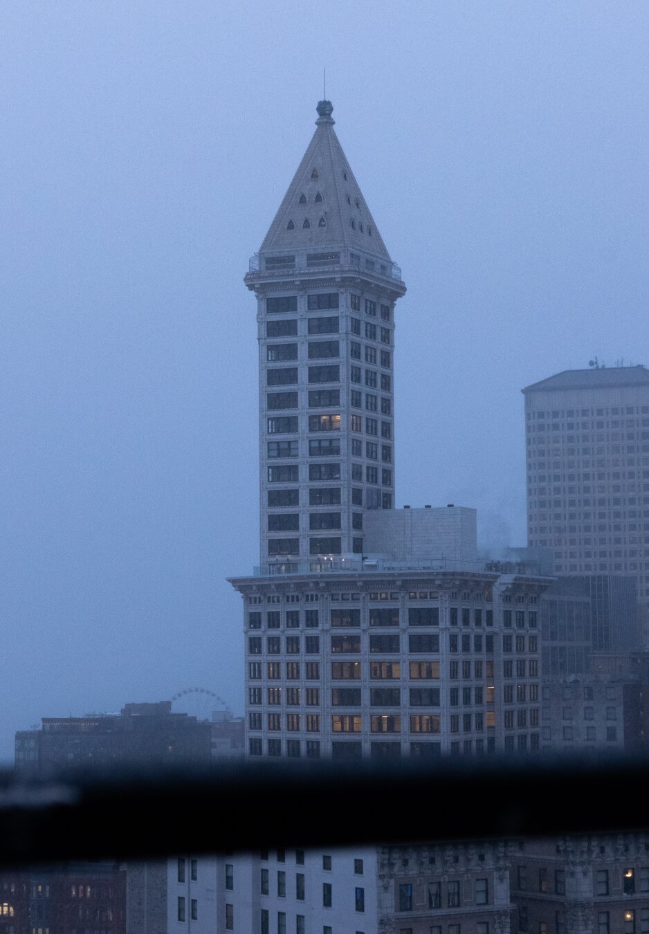 Portrait of Seattle's Smith Tower on a blue-grey, lightly snowy day. Behind it, low hanging fog covers nearby buildings. The photo is taken from a nearby rooftop with a guardrail at bottom frame, fuzzily out of focus.