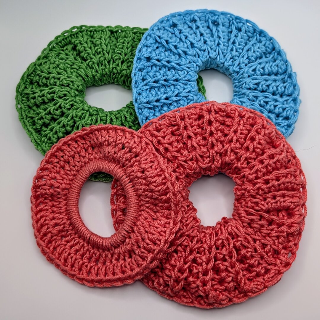 Four crocheted scrunchies stacked on each other in a vaguely square shape; one green, one blue, and two salmon. One of the salmon ones is smaller and a different style.